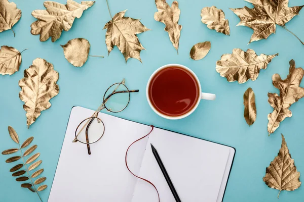 Draufsicht auf Tee in Tasse, Gläsern und Notizbuch bei goldenem Laub auf blauem Hintergrund — Stockfoto