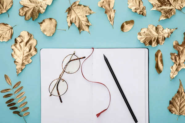 Top view of autumnal golden foliage near blank notebook with glasses on blue background — Stock Photo
