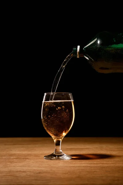 Cerveza vertiendo de botella en vidrio sobre mesa de madera aislada en negro - foto de stock