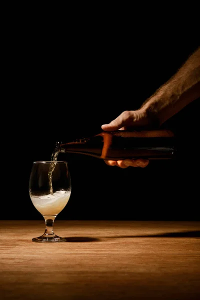Vue recadrée de l'homme versant de la bière de bouteille en verre sur table en bois isolé sur noir — Photo de stock