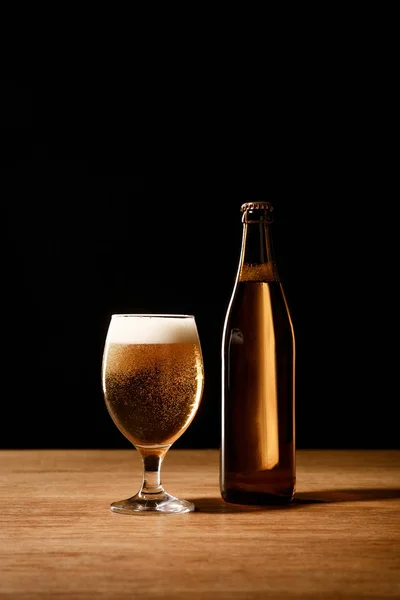 Cerveza en vidrio con espuma y en botella sobre mesa de madera aislada sobre negro - foto de stock