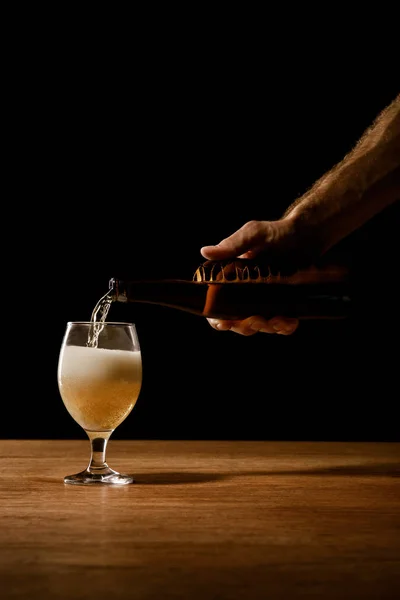 Visão parcial do homem derramando cerveja de garrafa em vidro sobre mesa de madeira isolada em preto — Fotografia de Stock