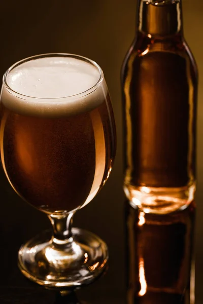 Vista de cerca de la cerveza con espuma en vidrio cerca de la botella sobre fondo oscuro con iluminación - foto de stock