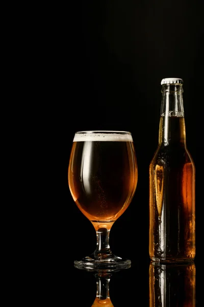 Vaso de cerveza con espuma cerca de botella aislada en negro - foto de stock