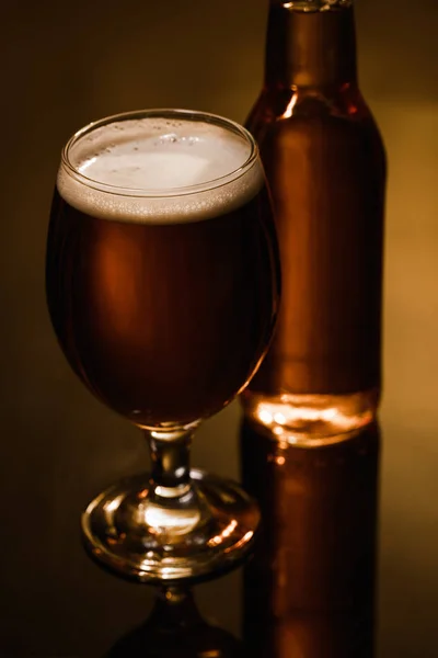 Selective focus of beer with foam in glass near bottle on dark background with lighting — Stock Photo