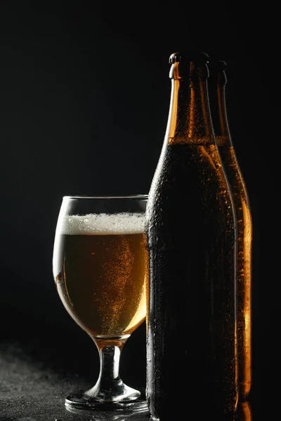 Botellas mojadas de cerveza con gotas cerca de vidrio con cerveza y espuma aislada en negro - foto de stock