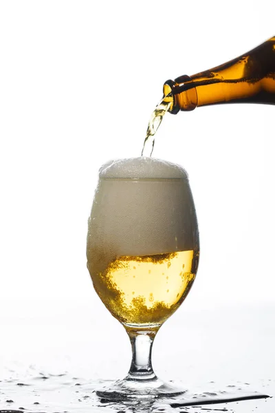 Beer pouring from bottle into glass on surface with puddle isolated on white — Stock Photo