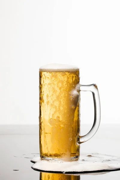 Wet glass of beer with foam and spilled puddle on surface isolated on white — Stock Photo