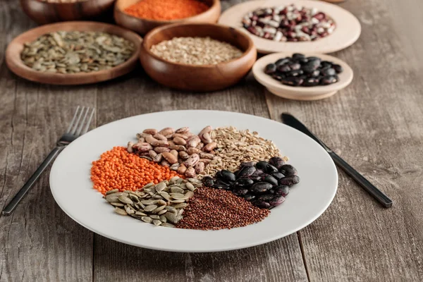 Plato blanco con lentejas crudas, frijoles, semillas de calabaza, quinua y avena con cuchillo y tenedor sobre mesa de madera - foto de stock