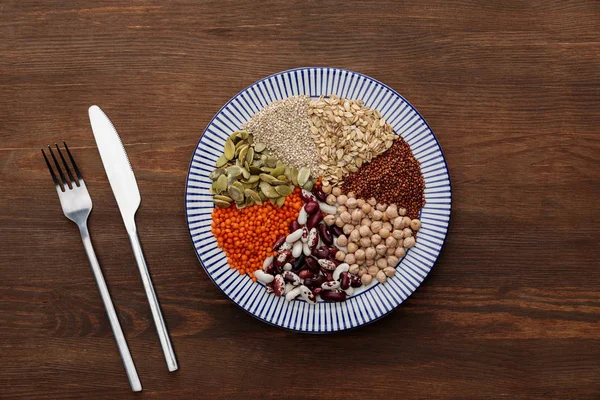 Blick von oben auf Gabel und Messer neben gestreiftem Teller mit rohen Linsen, Kichererbsen, Quinoa, Haferflocken, Bohnen und Kürbiskernen auf Holzoberfläche — Stock Photo