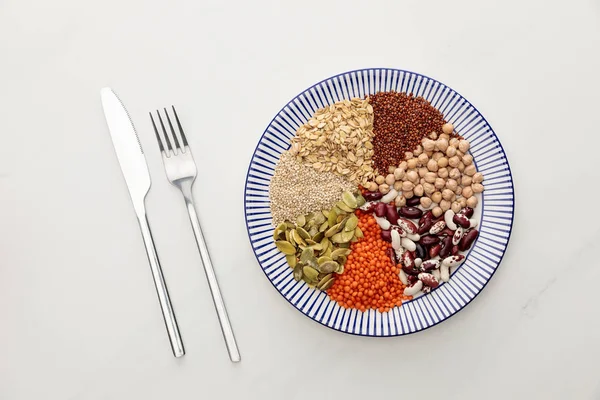 Blick von oben auf Gabel und Messer neben gestreiftem Teller mit rohen Linsen, Kichererbsen, Quinoa, Haferflocken, Bohnen und Kürbiskernen auf Marmoroberfläche — Stockfoto