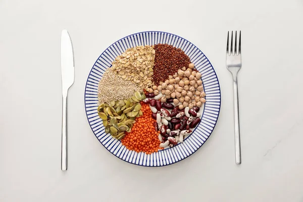 Top view of striped plate with raw lentil, chickpea, quinoa, oatmeal, beans and pumpkin seeds near cutlery on marble surface — Stock Photo
