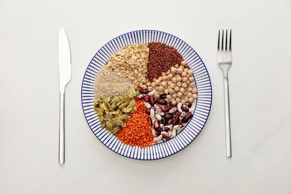 Vue de dessus de la plaque rayée avec lentille crue, pois chiche, quinoa, flocons d'avoine, haricots et graines de citrouille près des couverts sur la surface du marbre — Photo de stock