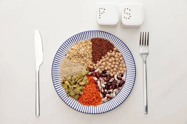 Vista dall'alto del piatto con cereali crudi, fagioli diversi e semi di zucca vicino a posate e pentole di sale e pepe sulla superficie di marmo — Foto stock