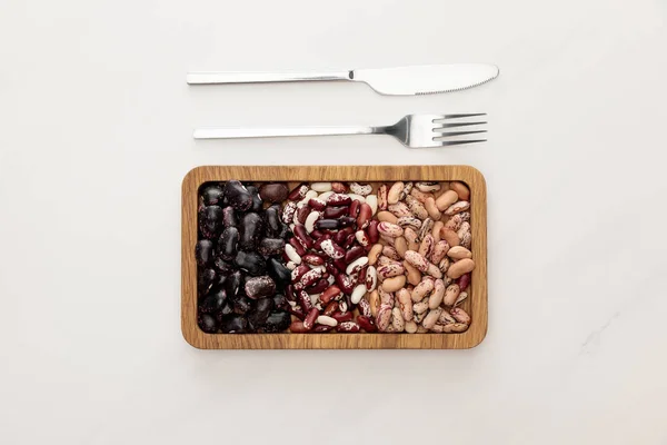 Vue de dessus du plat rectangulaire en bois avec haricots crus près de la fourchette et couteau sur la surface de marbre blanc — Photo de stock