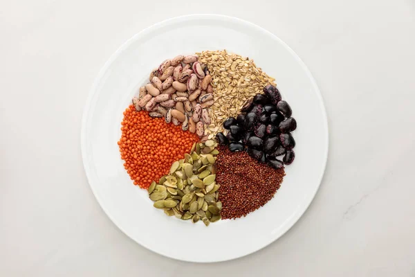 Vista superior da placa branca com lentilha crua, quinoa, aveia, feijão e sementes de abóbora na superfície de mármore — Fotografia de Stock