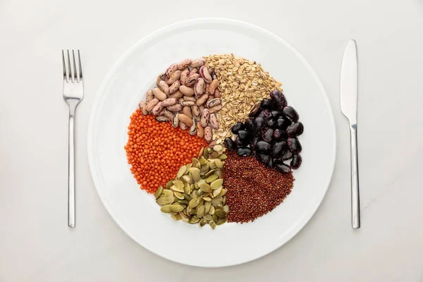 Top view of white plate with raw lentil, quinoa, oatmeal, beans and pumpkin seeds near fork and knife on marble surface — Stock Photo