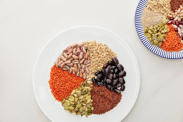 Vue de dessus des assiettes blanches et rayées avec lentilles crues, quinoa, flocons d'avoine, haricots et graines de citrouille sur la surface du marbre — Photo de stock