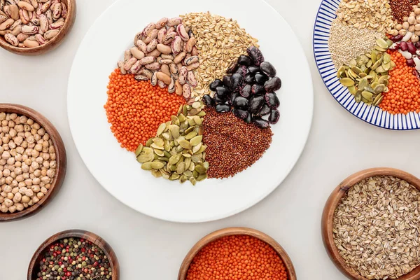 Vista dall'alto di piatti bianchi e rigati e ciotole di legno con lenticchie crude, quinoa, farina d'avena, fagioli, grani di pepe e semi di zucca sulla superficie di marmo — Foto stock