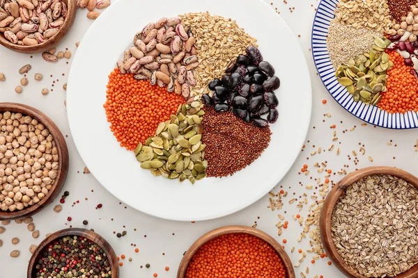 Top view of plates and bowls with raw lentil, quinoa, oatmeal, beans, peppercorns and pumpkin seeds on marble surface with scattered grains — Stock Photo