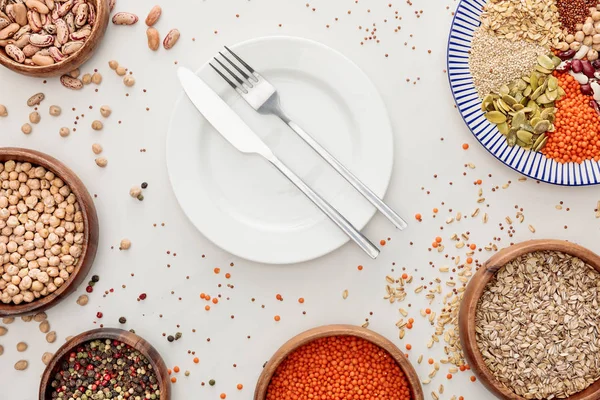 Vista superior de prato vazio com garfo e faca perto de tigelas com lentilha crua, quinoa, aveia, feijão, pimenta e sementes de abóbora na superfície de mármore com grãos espalhados — Fotografia de Stock