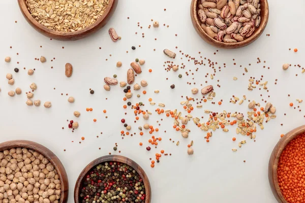 Vista superior de platos de madera con garbanzos, lentejas, granos de pimienta, avena y frijoles cerca de granos dispersos en la superficie de mármol - foto de stock