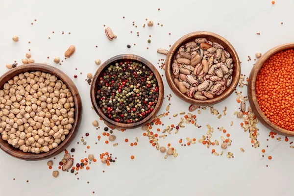 Top view of wooden plates with chickpea, lentil, peppercorns, and beans near scattered grains on marble surface — Stock Photo