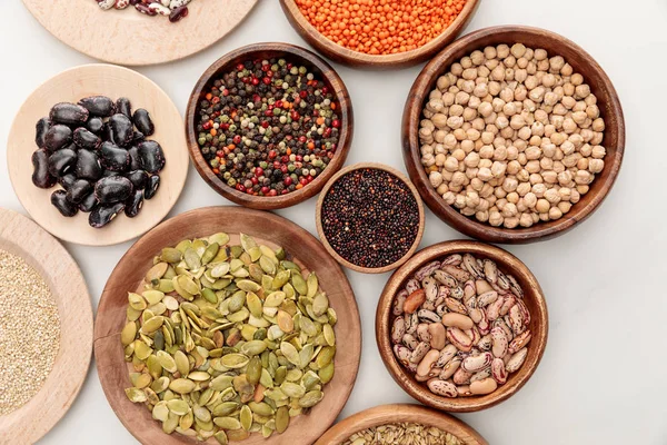 Vista dall'alto di ciotole di legno con fagioli diversi, quinoa, lenticchie rosse, grani di pepe, semi di zucca e ceci sulla superficie di marmo bianco — Foto stock