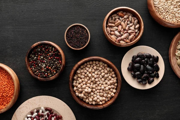 Top view of bowls with beans, chickpea, lentil, peppercorns, quinoa and oatmeal on dark wooden surface — Stock Photo