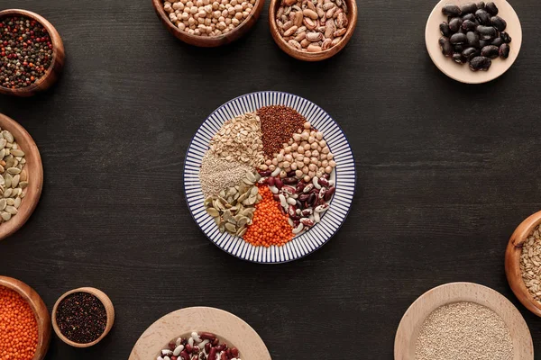 Vista dall'alto del piatto a strisce con vari legumi crudi e cereali vicino a ciotole su una superficie di legno scuro — Foto stock
