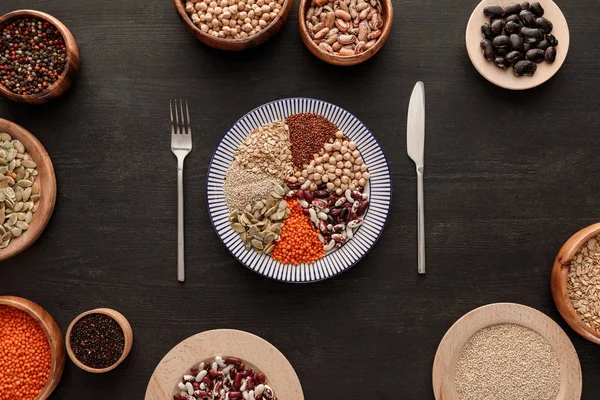 Top view of knife and fork near striped plate with various raw legumes and cereals and bowls on dark wooden surface — Stock Photo