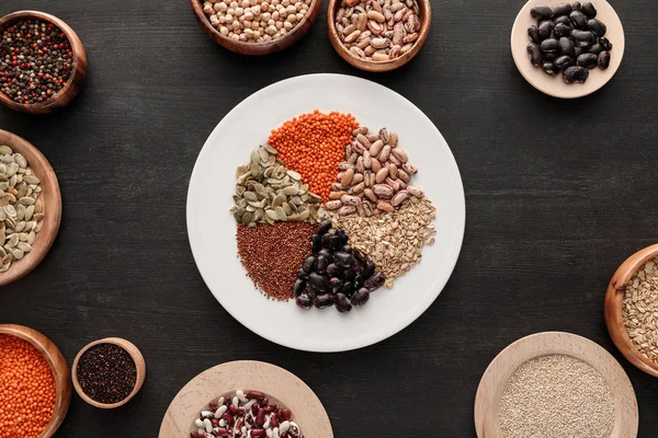 Top view of white plate with various raw legumes and cereals near bowls on dark wooden surface — Stock Photo