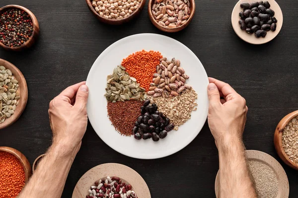 Cropped view of man holding white plate with various raw legumes and cereals near bowls on dark wooden surface — Stock Photo