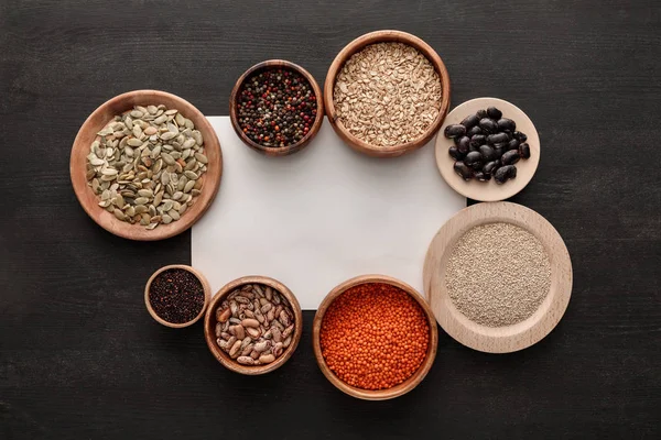 Vue du dessus du papier blanc vierge près de diverses assiettes et bols en bois avec haricots, céréales, épices et graines de citrouille sur table en bois sombre — Photo de stock