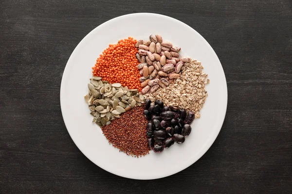 Top view of white ceramic plate with raw assorted beans, cereals and seeds on dark wooden surface with copy space — Stock Photo