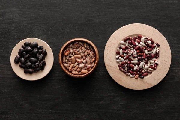 Vista superior de cuenco y platos con frijoles crudos surtidos en superficie de madera oscura con espacio para copiar - foto de stock