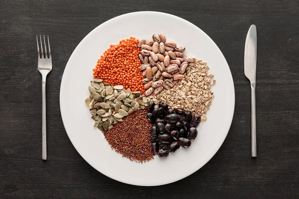 Top view of fork and knife near white ceramic plate with raw assorted beans, cereals and seeds on dark wooden surface with copy space — Stock Photo