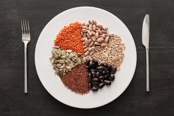 Vista superior de tenedor y cuchillo cerca de placa de cerámica blanca con frijoles crudos surtidos, cereales y semillas en la superficie de madera oscura con espacio para copiar - foto de stock