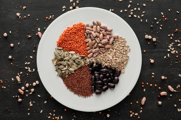 Top view of white ceramic plate with raw assorted beans, cereals and seeds on dark wooden surface with scattered grains — Stock Photo