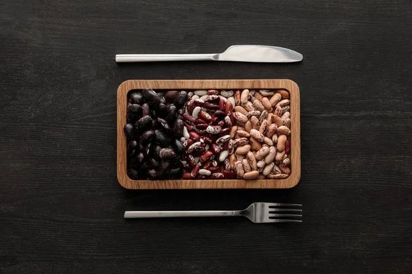 Top view of fork and knife near rectangular brown bowl with raw assorted beans on dark wooden surface — Stock Photo
