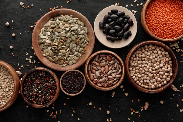 Top view of wooden bowls and plates with raw assorted beans, cereals and seeds on dark surface with scattered grains — Stock Photo