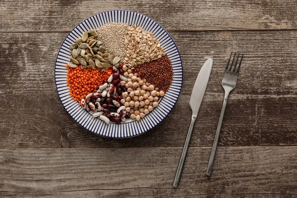 Vista superior de tenedor y cuchillo cerca de placa de cerámica a rayas con frijoles crudos surtidos, cereales y semillas en la superficie de madera oscura con espacio de copia - foto de stock