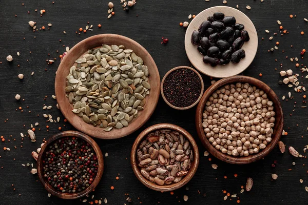 Top view of wooden bowls and plates with raw beans, chickpea, quinoa, pumpkin seeds and peppercorns on dark surface with scattered grains — Stock Photo