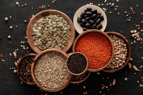 Vista superior de cuencos y platos de madera con lentejas crudas, harina de avena, frijoles, garbanzos, quinua, semillas de calabaza y granos de pimienta en la superficie oscura con granos dispersos - foto de stock