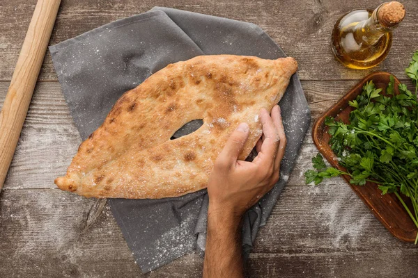 Cropped view of man holding lavash bread near rolling pin, fresh parsley and olive oil on wooden table — Stock Photo