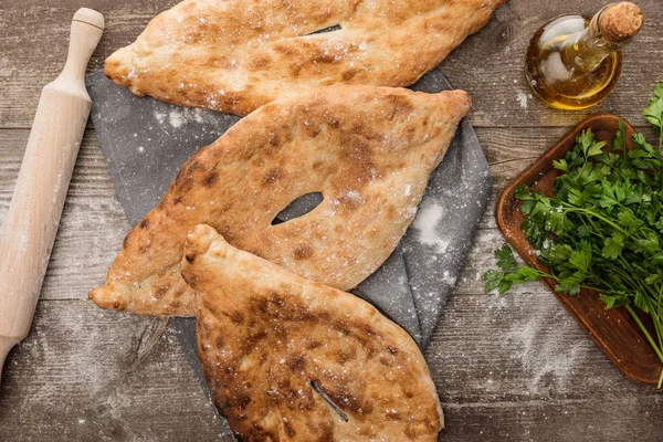 Vista dall'alto del pane lavash su asciugamano grigio vicino al mattarello, prezzemolo fresco e olio d'oliva sul tavolo di legno con farina sparsa — Foto stock