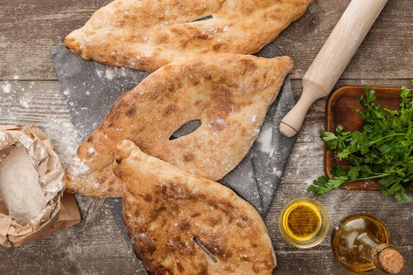 Vue du dessus du pain de lavande sur une serviette grise près du rouleau à pâtisserie, paquet avec farine, persil frais et huile d'olive sur table en bois — Photo de stock