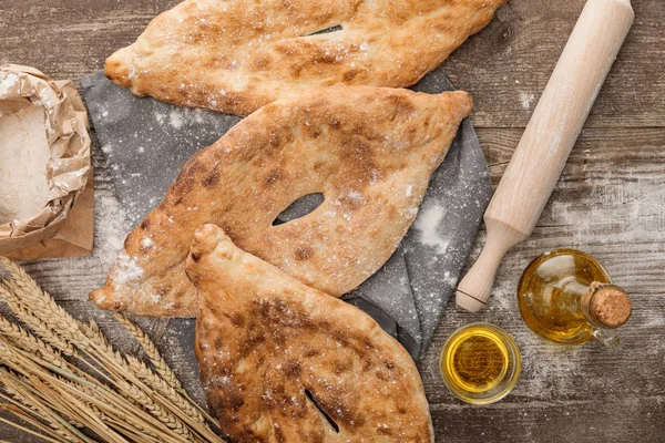Vue du dessus du pain de lavande sur serviette près du rouleau à pâtisserie, paquet avec farine, huile d'olive et épis de blé sur table en bois — Photo de stock