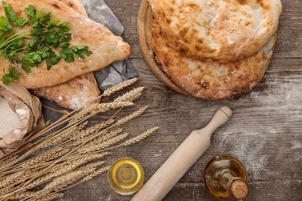 Top view of lavash bread on gray towel near flour package, wheat spikes, fresh greenery and olive oil on wooden table — Stock Photo