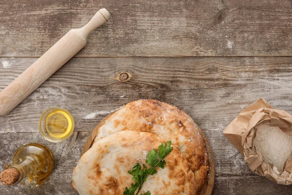 Vue du dessus du pain de lavande près du paquet de farine, rouleau à pâtisserie, persil frais et bouteille d'huile d'olive sur table en bois — Photo de stock